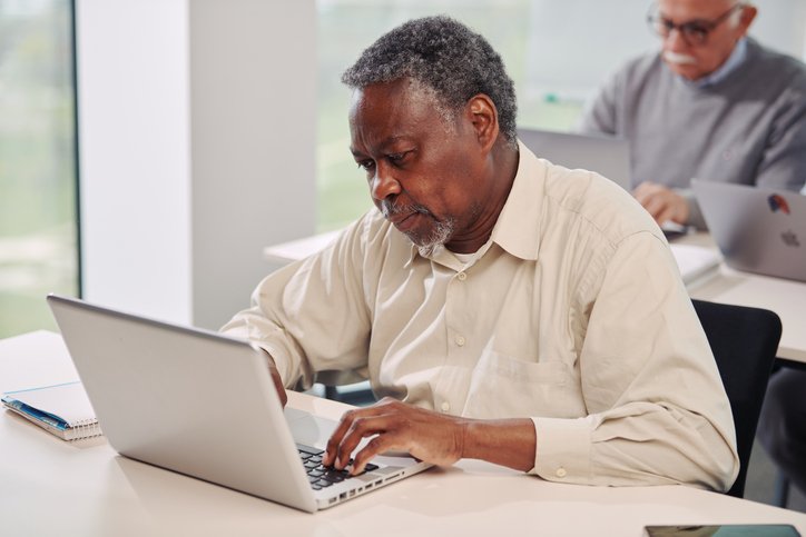 A multicultural senior student sits in a classroom and having an exam on computer with his classmates.